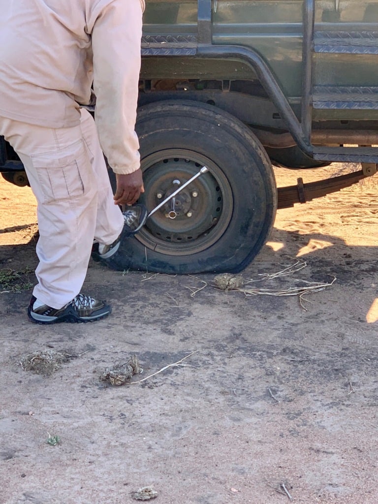 Someone in khaki pants crouching down to work on a flat tire on the safari vehicle in Kruger, red dirt and scattered branches on the ground around them.