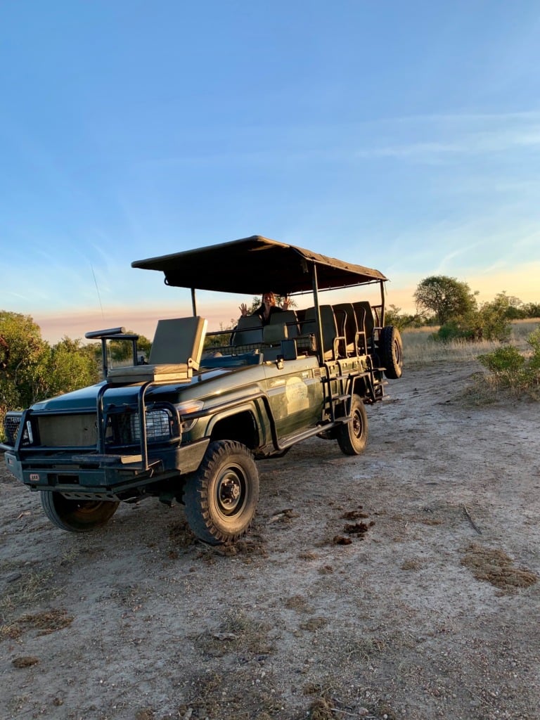 An open-sided safari truck parked on dusty ground in Kruger at golden hour, the sky turning orange behind it, grassland stretching out in all directions.
