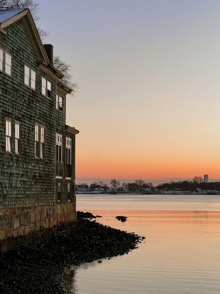 A weathered green-shingled building on the edge of Portsmouth Harbor in New Hampshire at sunset, with the water glowing orange and the far shore visible across the calm harbor