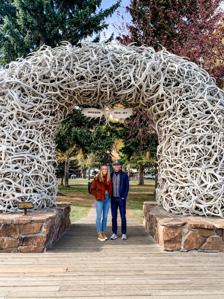 A couple standing beneath the iconic elk antler arch at Jackson Hole Town Square in Wyoming, surrounded by fall foliage