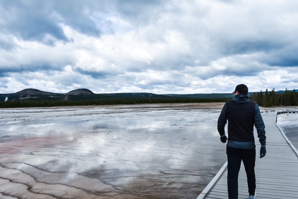 A person standing on a boardwalk overlooking a vast thermal basin in Yellowstone National Park, with steam rising from the ground and mountains in the distance