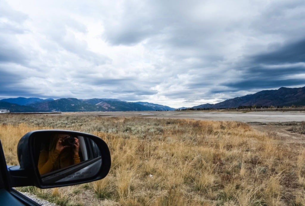 A photographer's reflection in a car side mirror while shooting the wide sage and grassland landscape on a road trip through Jackson Hole, Wyoming