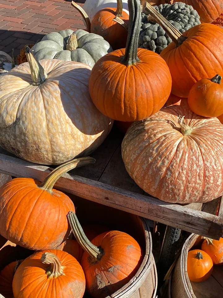 A wooden cart piled with orange, white, and sage green pumpkins of various sizes at a fall harvest market, with cobblestone pavement in the background